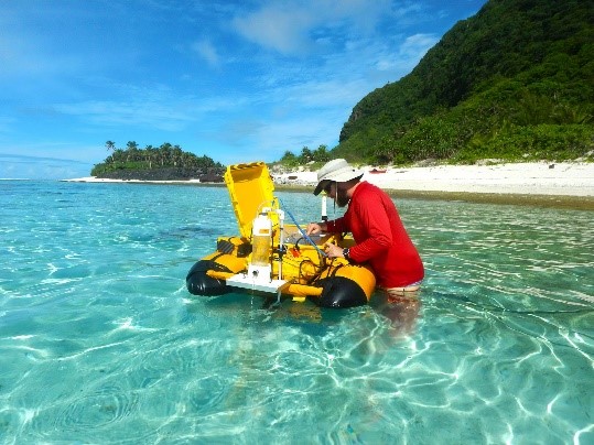 A photo of a tropical looking ocean, beach in background, clear water and blue skies. In the center of the image is a man wearing a red UV shirt. He is working on a large orange autonomous vehicle used for marine remote sensing.
