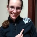Headshot of Elizabeth, a white woman with brown hair in a ponytail, wearing glasses. She is wearing a black jacket and has a gray 3D printed trilobite on her shoulder, which she is pointing to.