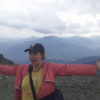 Photo of Jana, a white woman wearing a baseball hat and a pink jacket on a mountain in Flims, Switzerland. Her arms are outstretched and she is grinning, with clouds and mountains in the background.