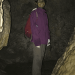Natasha is a brown-skinned woman, wearing glasses and in a pink rain jacket, brown pants with a red backpack. She is wearing a cave helmet with two headlamps and nitrile gloves. She is standing inside a cave in southeastern New Mexico.