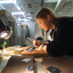 Chrissy is a white woman wearing a dark jacket. She is in a museum collections room painting liquid latex onto fossils, using a lamp to help them dry.