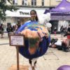 Rehemat is a brown-skinned woman, wearing a globe costume depicting the Earth. Her hair is tied back in a ponytail, and she is standing on a wooden box in a pedestrianised shopping area