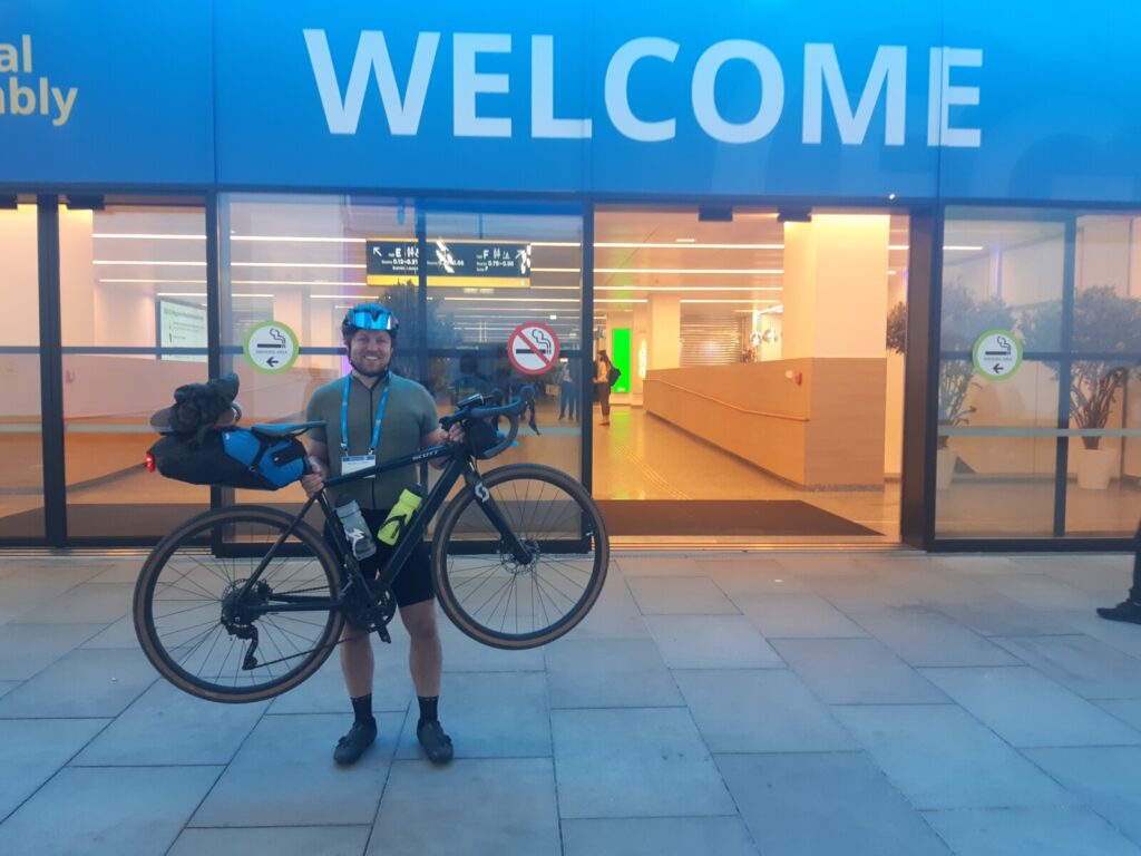 A happy ben lifting up his bike in front of the conference center under a sign saying 'Welcome'