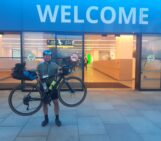 A happy ben lifting up his bike in front of the conference center under a sign saying 'Welcome'