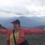 Photo of Jana, a white woman wearing a baseball hat and a pink jacket on a mountain in Flims, Switzerland. Her arms are outstretched and she is grinning, with clouds and mountains in the background.