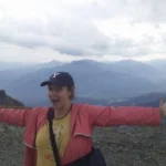 Photo of Jana, a white woman wearing a baseball hat and a pink jacket on a mountain in Flims, Switzerland. Her arms are outstretched and she is grinning, with clouds and mountains in the background.
