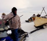 Man on snow machine looks bake to the canoe he is towing across ice.