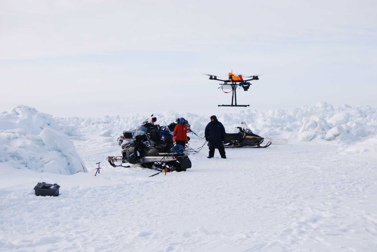 A drone is lifting up in front of people and snow machines in a wide snow landscape