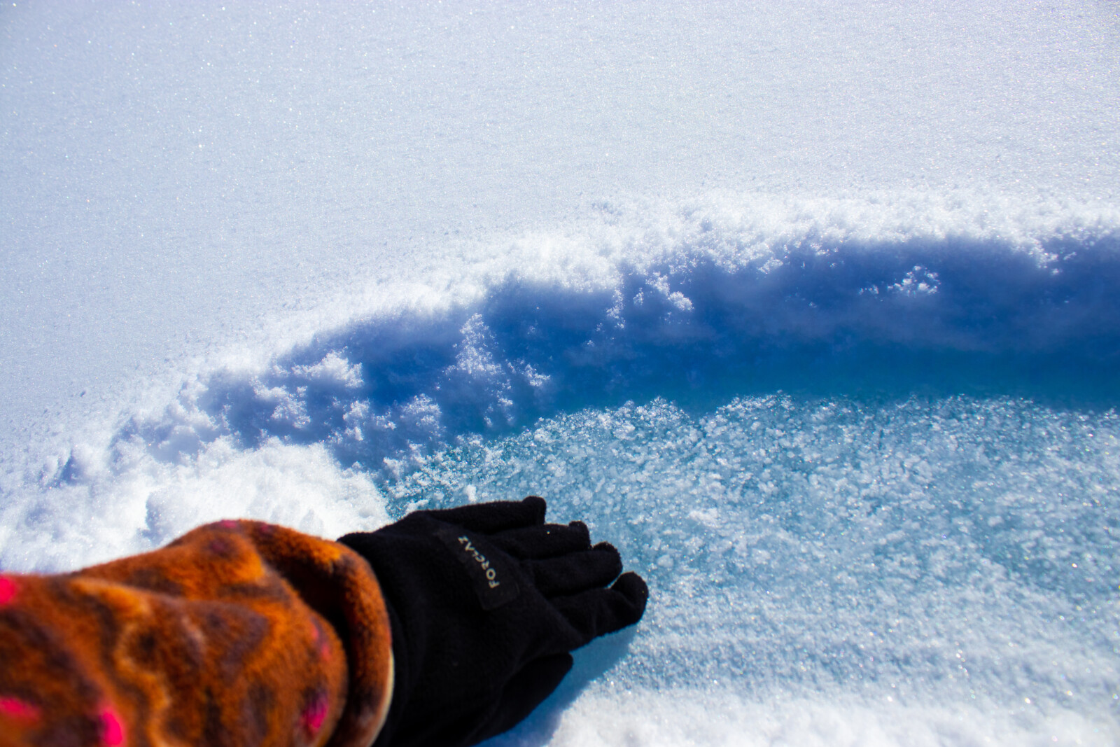 A gloves hand swipes away snow to find blue ice underneath.