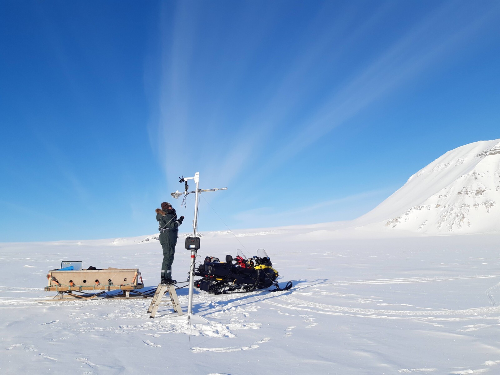 a female field scientist stood on a ladder in the snow installing a weather station, with snow mobiles behind her.