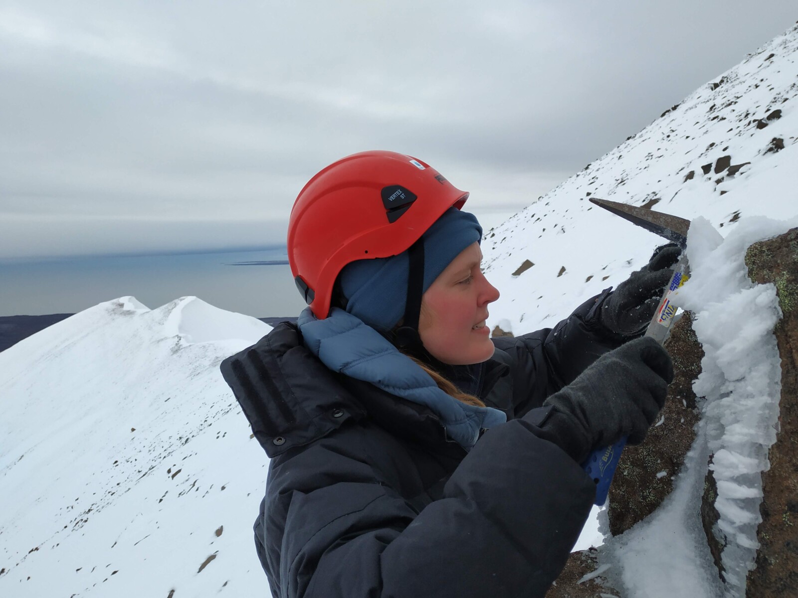 a female scientists using an axe to take rock samples from a rocky outcrop in a snowy landscape.