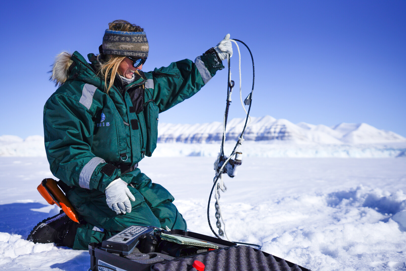 A female scientists holding up cables on top of sea ice with snowy mountains in the back ground, and an open box with scientific equipment in front of them. 