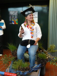 A person with a hat on top and a bottle in the hand sits on a trolley decorated with green twigs.