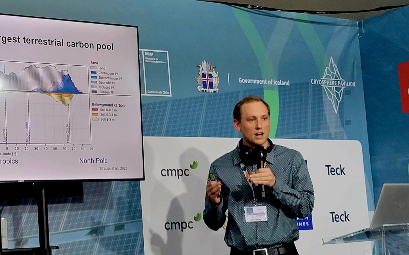 Author stood to the right of a poster board holding a microphone, giving a scientific presentation.