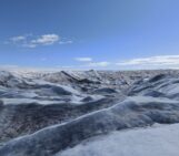 Below a clear blue sky, ridges of ice undulate towards the horizon in stripes of white, blue, and black.