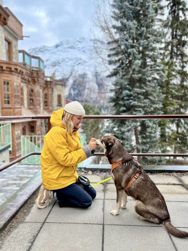 a young woman with blonde hair and dressed for cold waeather, sits with a dog in a swiss city, in front of a mountain landscape