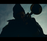 against a blue, muted sky a saxophone layer in a beanie plays a song