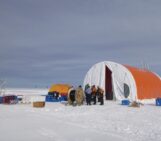 an orange, rugged large tent is in an arctic landscape. researchers in cold-eather gear gather at the mouth of the tent.