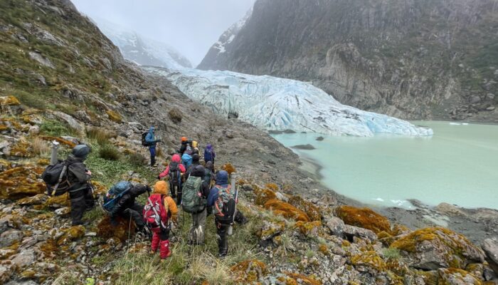 a group of students and staff in hi-vis rain gear pick theri way along a rocky slope towards a glacier