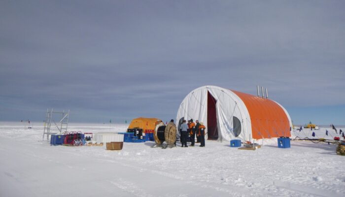 an orange, rugged large tent is in an arctic landscape. researchers in cold-eather gear gather at the mouth of the tent.