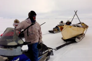 Man on snow machine looks bake to the canoe he is towing across ice.
