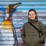 Woman in khaki jacket besides a penguin graffiti.