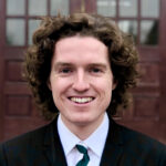 Person with curly hair, smile and black and white suit and tie.