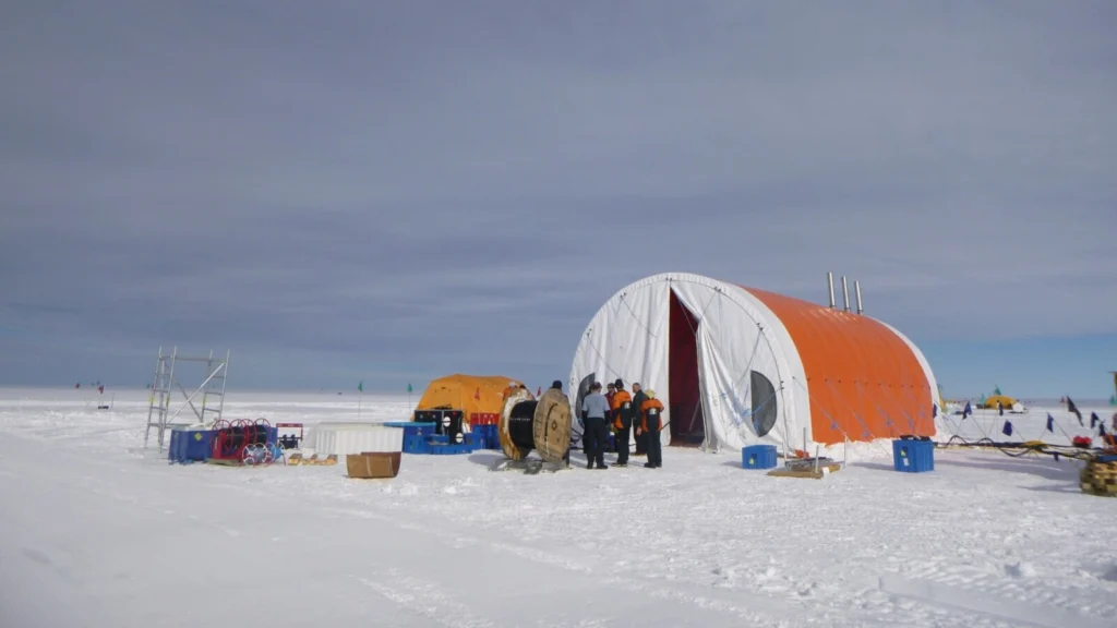 an orange, rugged large tent is in an arctic landscape. researchers in cold-eather gear gather at the mouth of the tent.