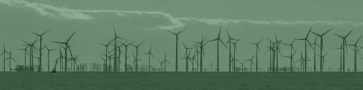 Tinted detail of a photo: Rows of wind turbines silhouetted on the horizon against a golden sunset band beneath dark clouds, seen across calm grey sea and wet sand.