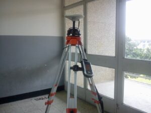 A white GPS antenna on a silver tripod inside a building in front of a large window.