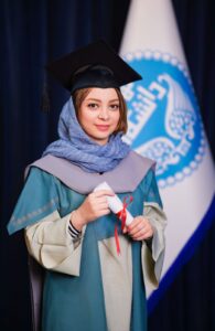 Kiana in a long blue and beige dress and a blue hijab covered by a black graduation hat. She smiles lightly and holds a white paper role hold together by red cord.