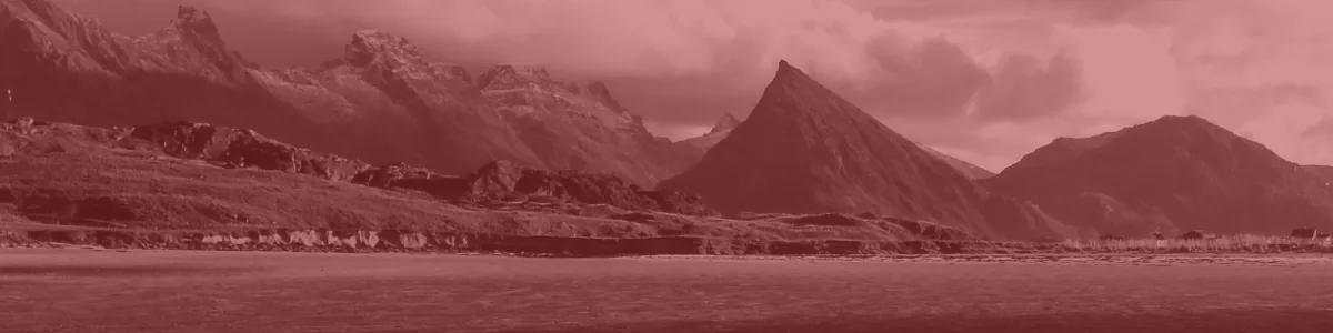Tinted detail of a photo: Wet sand on a beach rippled into many small wavy ridges, with a shallow channel of water behind and grassy land below sharp, dark, partly snow-dusted peaks under white clouds.