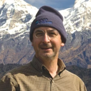 a portrait of Christian France Lanord in the field, with snow-capped mountains in the background and Christian in the foreground. He wears a brown shirt and a navy beanie hat. 