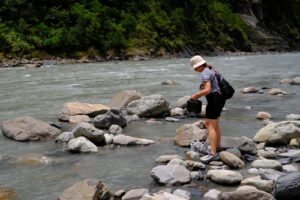 A person stands on rocks at the edge of a river holding a sampling box. The river water is grey with suspended particles. 