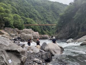 3 people stand and sit on rocks at the edge of a river, looking downstream. In the distance there's a bridge across the river, between two steep cliffs/hills covered in vegetation. 