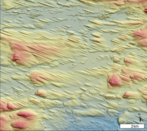 An aerial/satellite view topography (with low topography in blue, grading through greens and yellows, into higher topography coloured red). The topography illustrates linear features of topography ("Mega Scale Glacial Lineations (MSGL)") that all run parallel to one another, from West-North-West to East-South-East. Image is approximately 10 km by 10 km