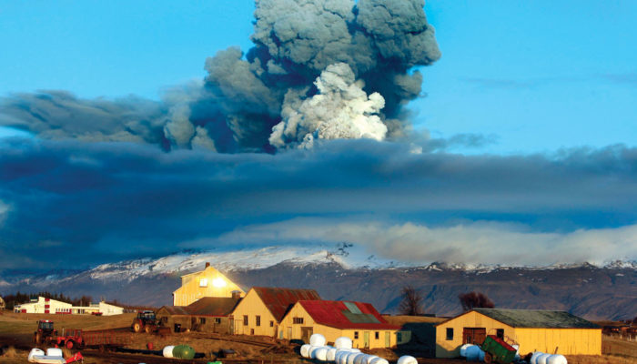In the foreground there is a farm with a backdrop of a volcano is emitting large amounts of ash into the air.