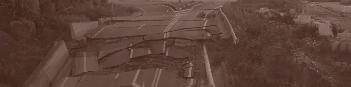 Tinted detail of a photo: Road broken into slabs and buckled chunks of asphalt amid heaped soil, with a bent crash barrier and damaged buildings beside it, leading to a roundabout under a hazy setting sun.
