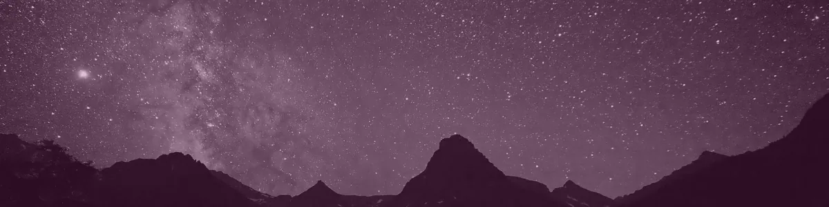 Tinted detail of a photo: Night sky full of stars with the Milky Way rising in a pale band above dark, pointed mountain silhouettes, mirrored in a still lake where the stars appear as short streaks.