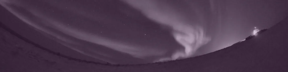 Tinted detail of a photo: Fisheye view of bright green aurora bands swirling across a starry night sky above a snowy curved horizon, with a small lit building at the right edge.