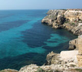 an image of a sea cove with rugged rocks to the right and bottom right of the image and clear blue sea water to the left and upper left of the image