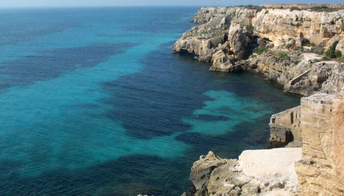 an image of a sea cove with rugged rocks to the right and bottom right of the image and clear blue sea water to the left and upper left of the image