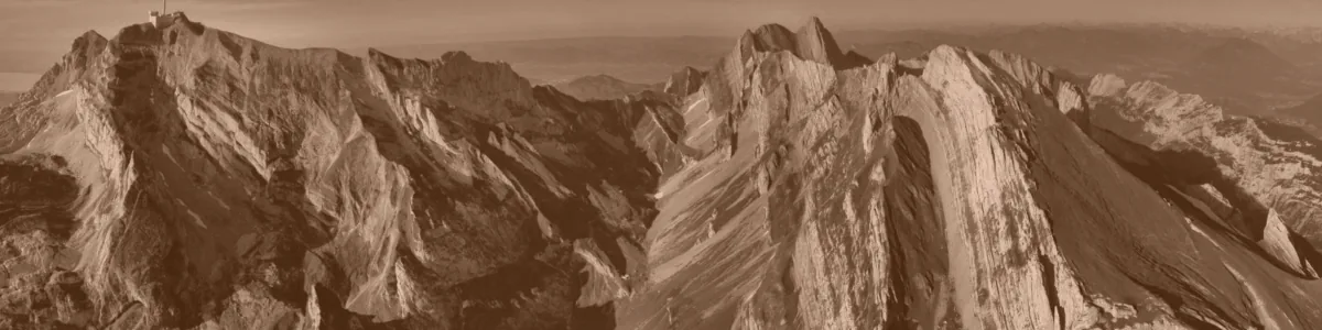 Tinted detail of a photo: Aerial view of steep limestone mountain ridges whose pale rock layers are tilted almost vertical, with green grassy slopes between them, a deep shadowed valley and a building with a mast on the left summit.