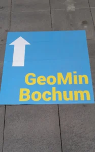 Blue Square on the ground with a white arrow pointing forward and a yellow writing say "GeoMinBochum"