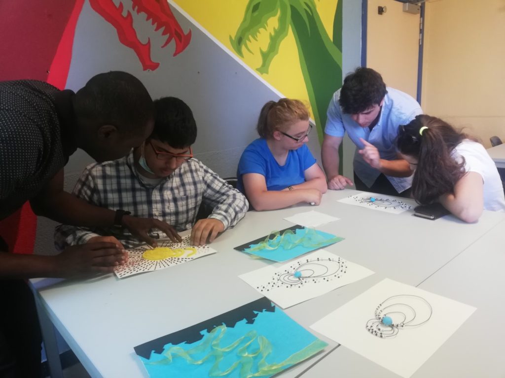 Blind and visually impaired students interact with the tactile images of space weather, assisted by teachers and researchers. Several tactile images are spread on the tables in from of three students, as they feel the phenomena being described by the images.