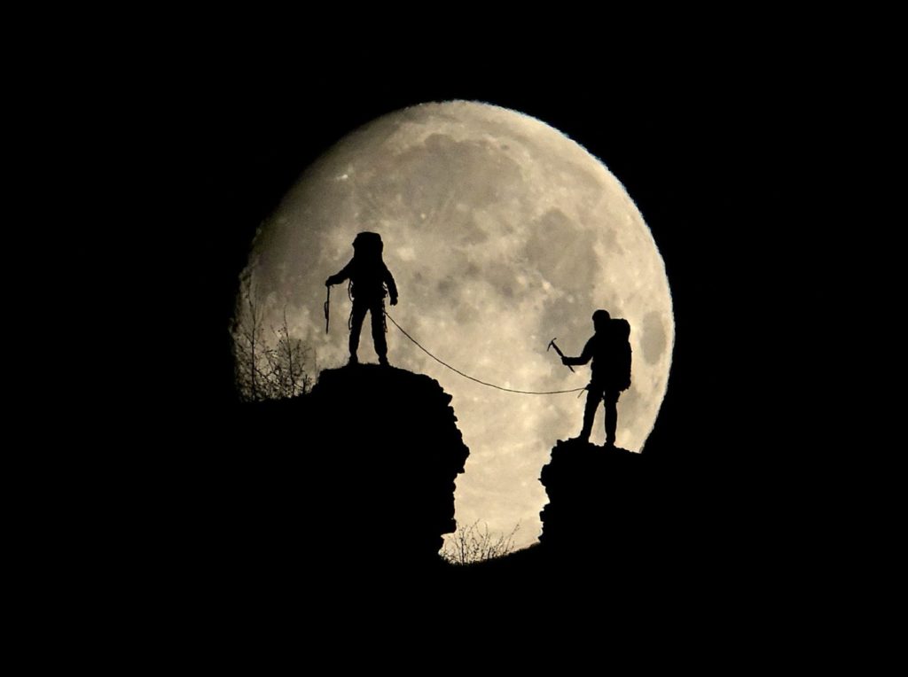 silhouette of two hikers in front of moon by Cyril Mayaud