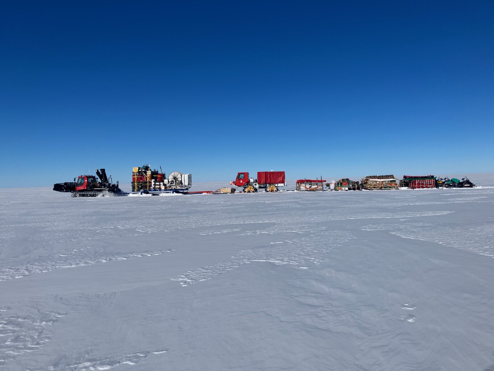 A train of different vehicles and tents on a wide white horizon with expansive blue skies.