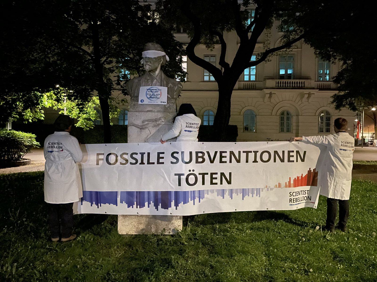 At night, two people dressed in lab coats hold a banner in front a blindfolded statue. The banner says in German that fossil subsidies kill.