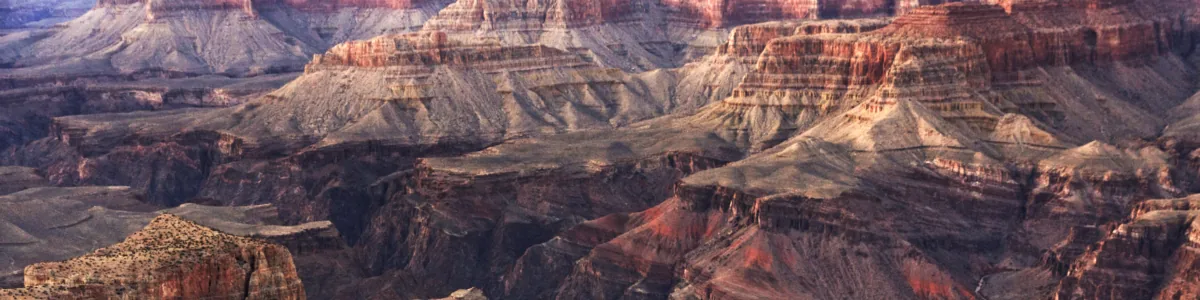Detail of a photo: Maze of layered canyon walls and buttes in bands of red, cream and grey, receding into the distance in soft evening light.