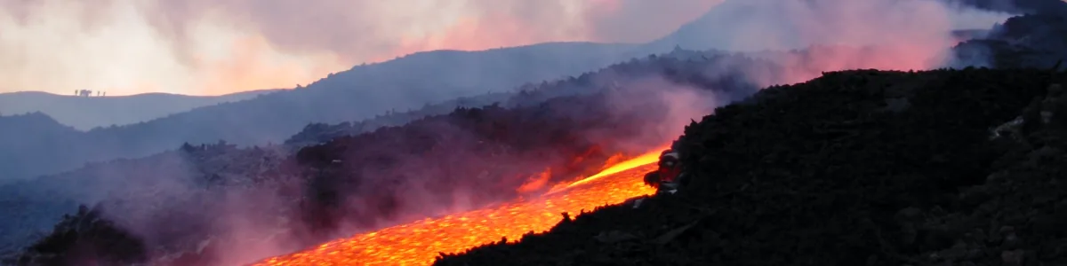 Detail of a photo: Glowing orange lava flow running down a dark slope at twilight, with smoke and pink-lit clouds rising past a dark cone-shaped hill behind it.